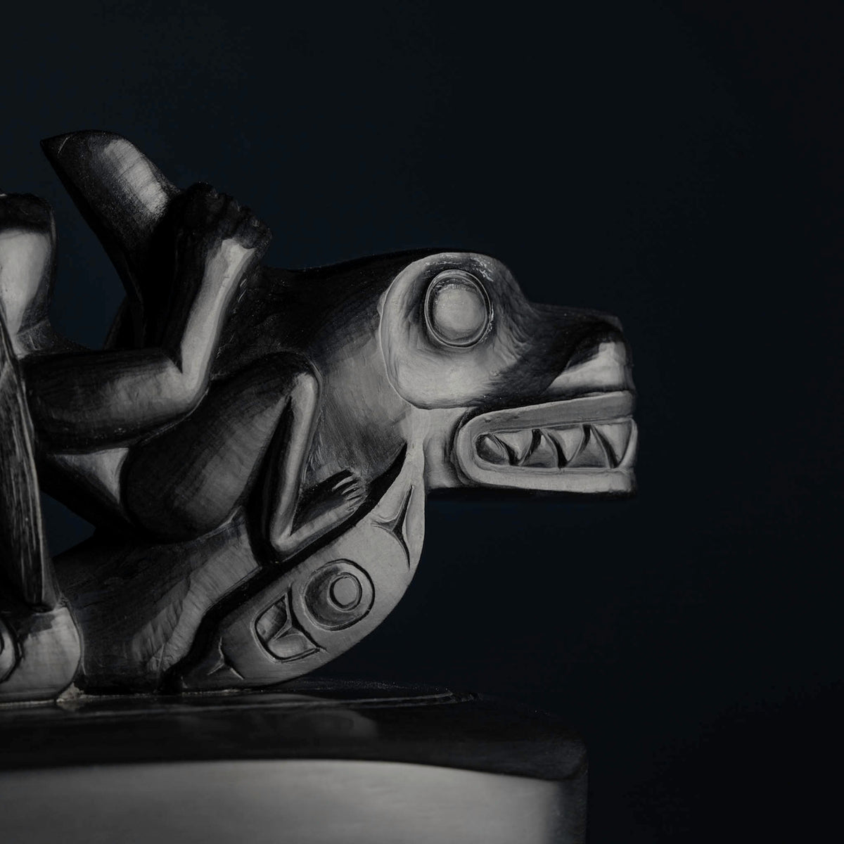 A close-up of a dark carved argillite depicting a killer whale with a person on its back, set against a very dark background.