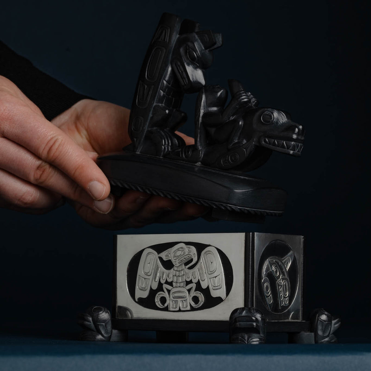 A hand lifts up the lid of an argillite box with a dark background.