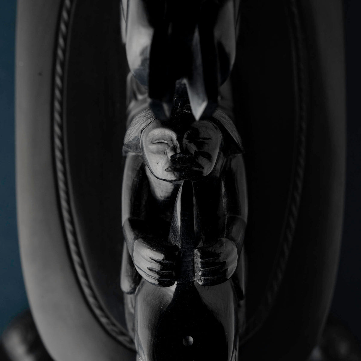 human face carved in black stone holing an object in their hands with rope like carvings on the black stone below.