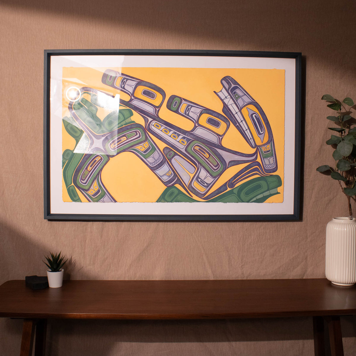 Framed abstract artwork on a wall with a wooden table and plant in the foreground.