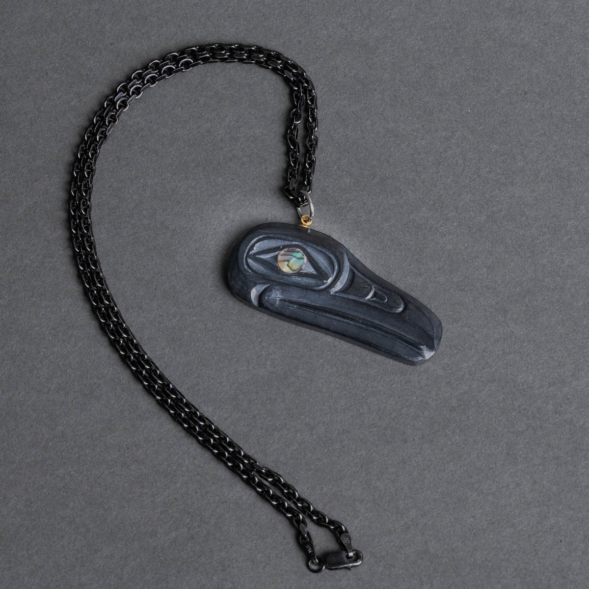 A dark carved raven pendant with a small iridescent eye detail, on a black chain on a textured gray background.