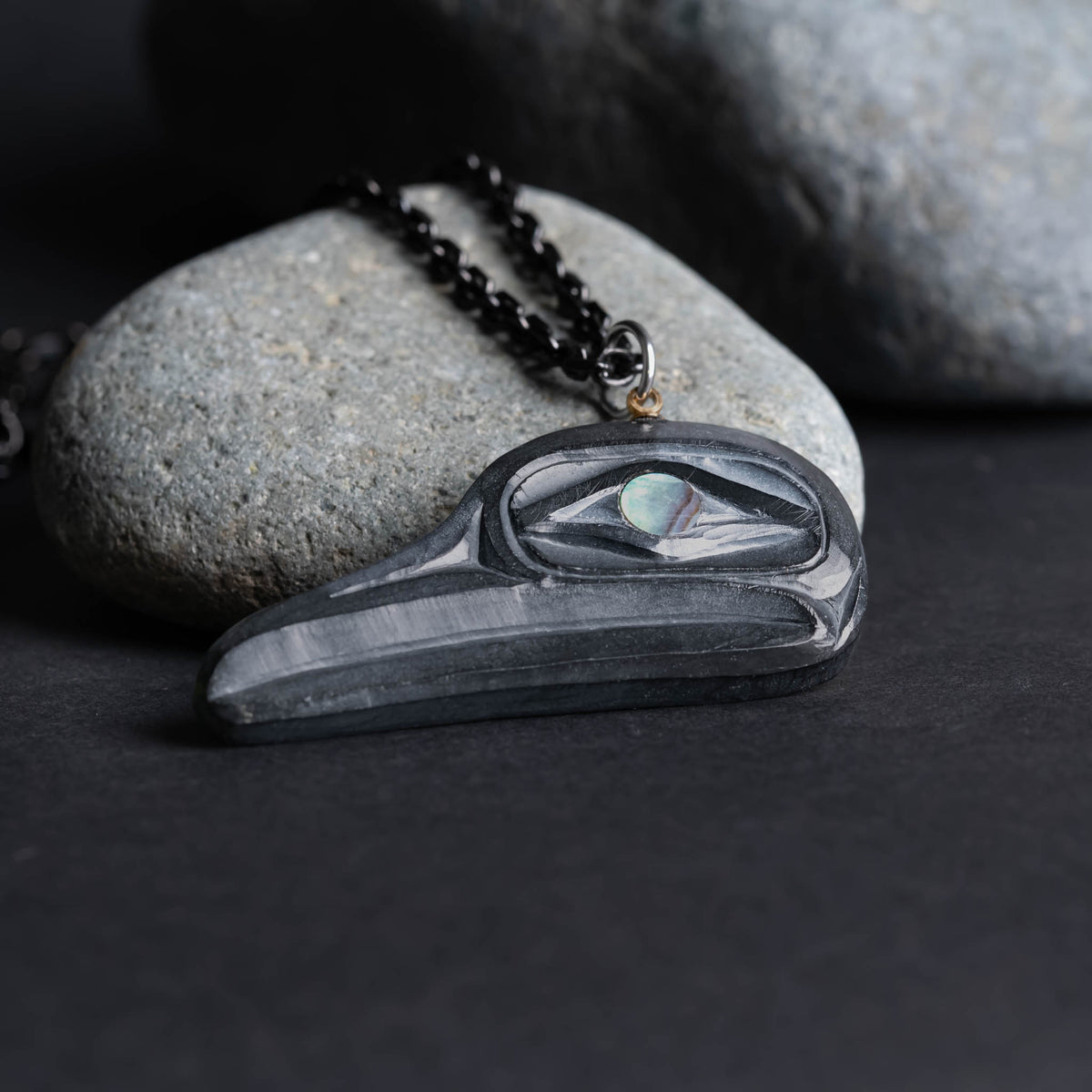 Close up of argillite flicker pendant showing its depth, proped against a ight grey rock on a dark grey background.