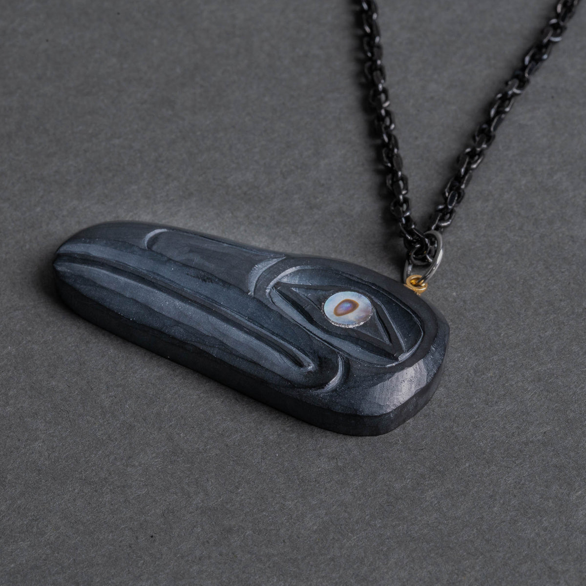 A photo of a Haida argillite raven pendant facing left with an abalone eye and black chain pointing to the top right corner.