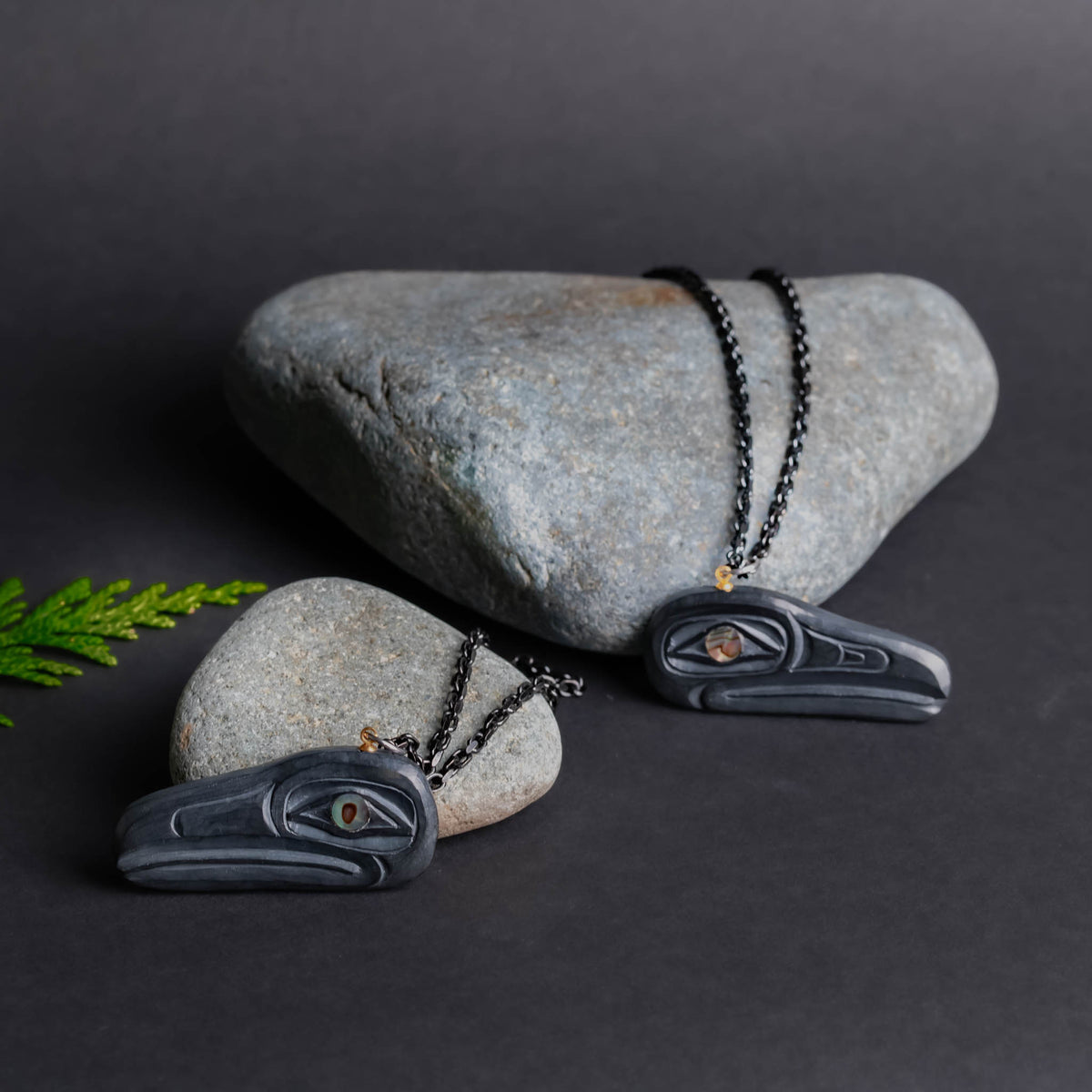 Two dark grey stone necklaces proped against light grey rocks with a cedar sprig beside them, one a dork grey backgound.