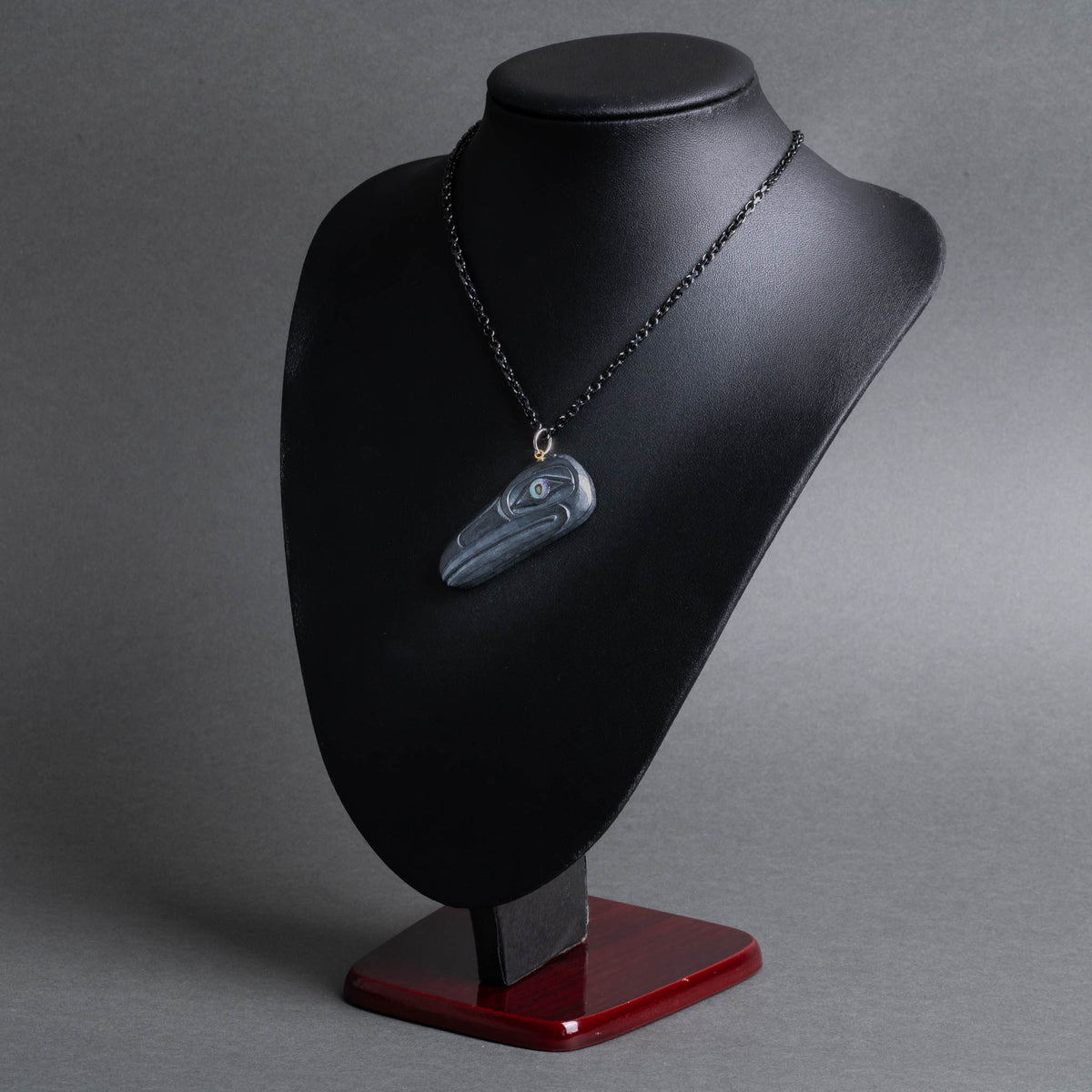 A carved stone raven necklace displayed on a black jewelry bust with a dark red base, photographed against a smooth gray studio background.