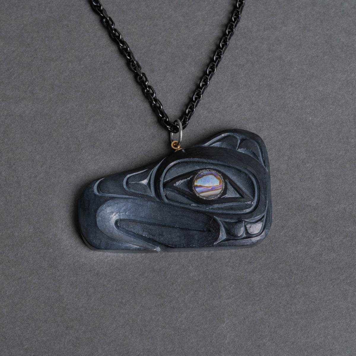 A carved dark stone pendant necklace with a thunderbird design and a small iridescent eye detail, hanging from a black chain on a dark gray background.