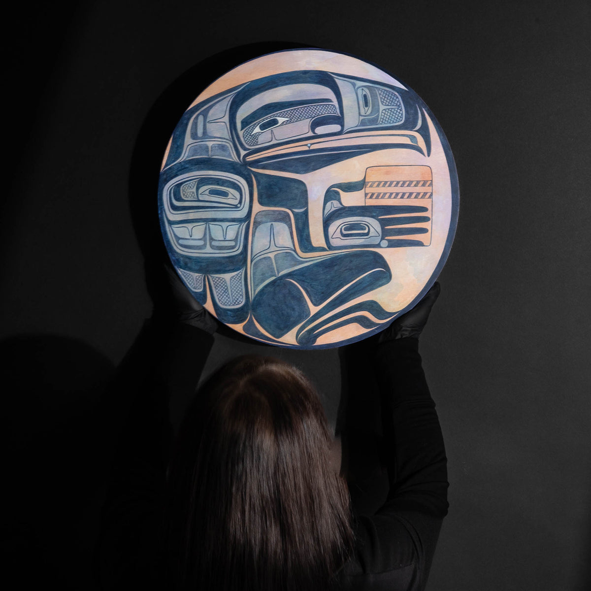 Person holding a round art piece with intricate blue and orange design against a dark background.