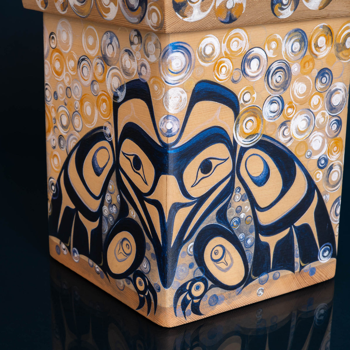 A close-up of a decorative square box with a painted Indigenous-style bird face and circular patterns in navy, gold, white, and tan on a dark reflective surface.