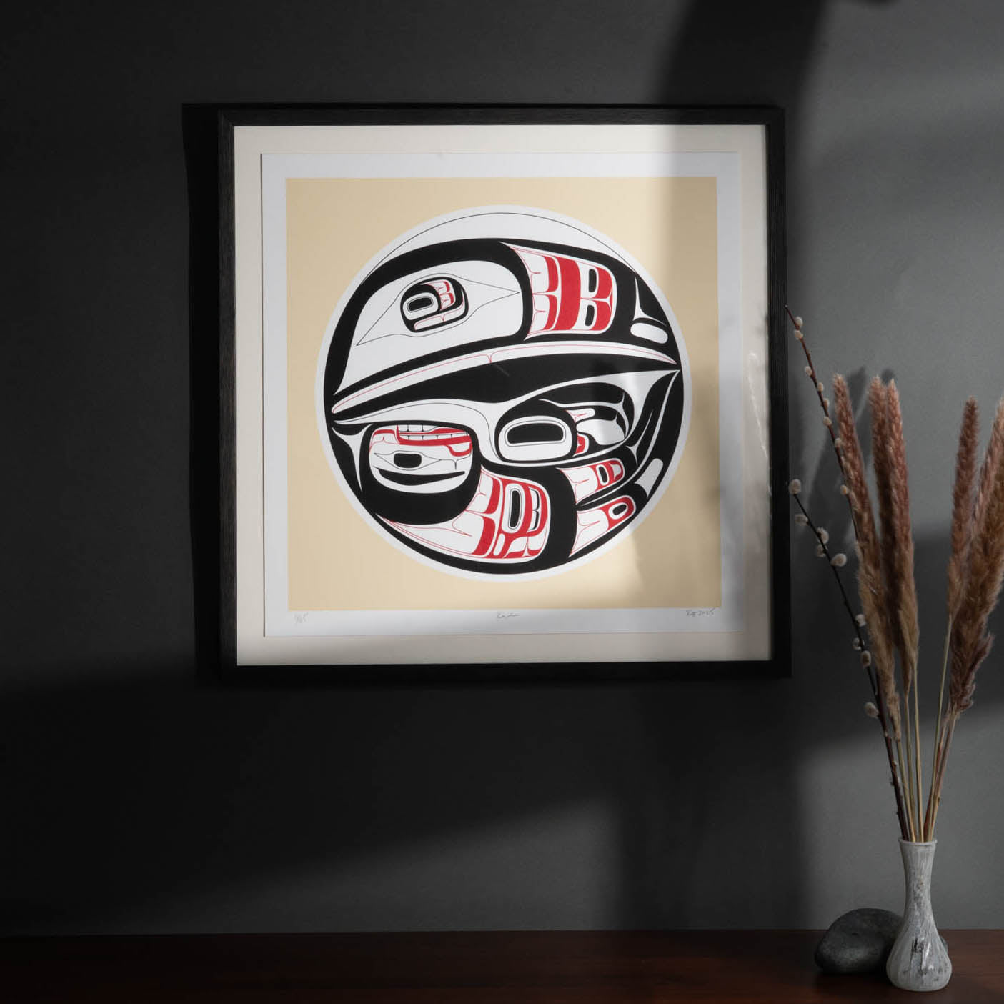Framed artwork with a stylized formline raven design on a beige background, placed on a dark surface.