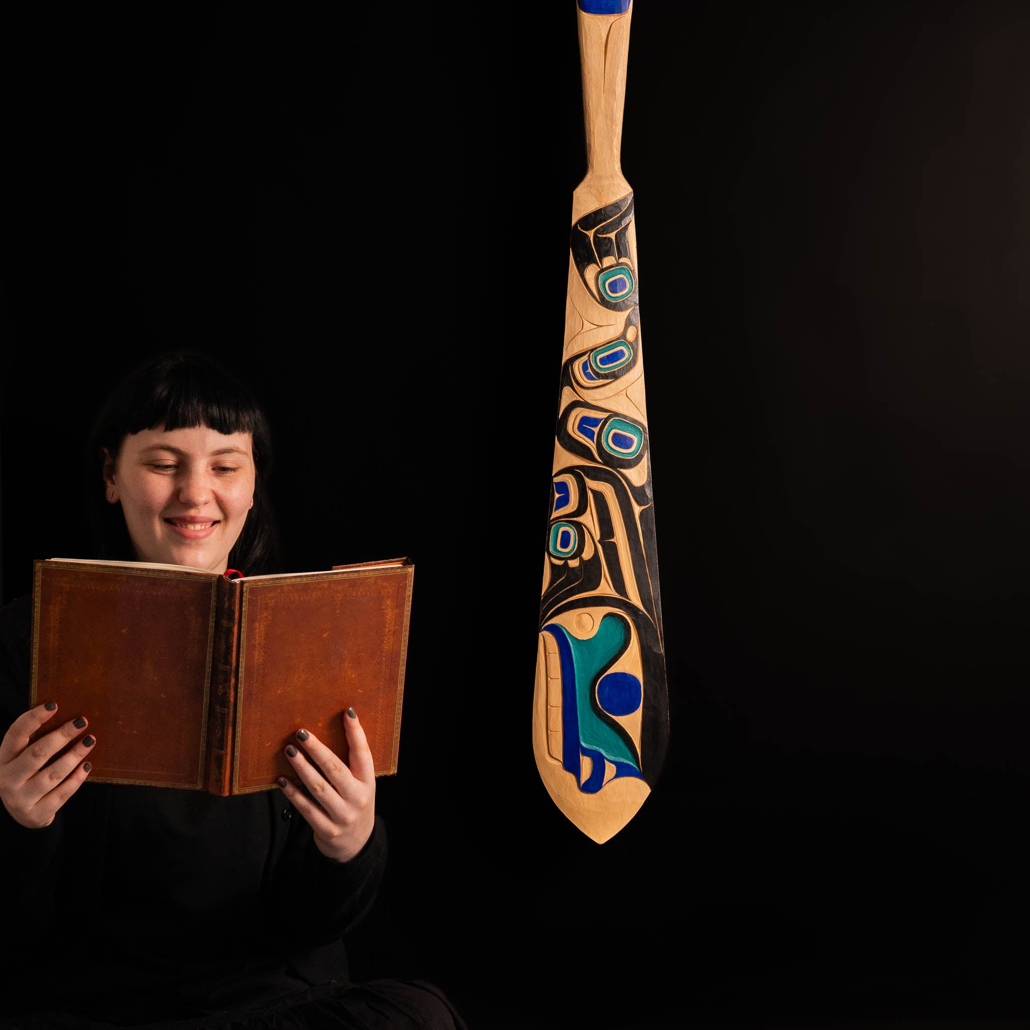 Person holding a book next to a large, intricately designed wooden paddle on a black background