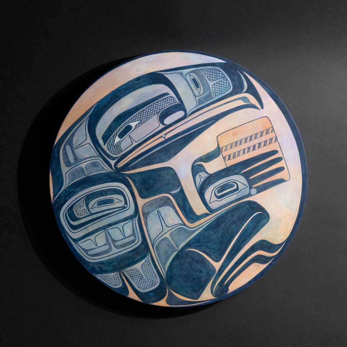 Round hand-painted original Haida painting featuring a blue raven in a pink and orange moon photographed on a black background by Haida artist Shoshannah Greene.