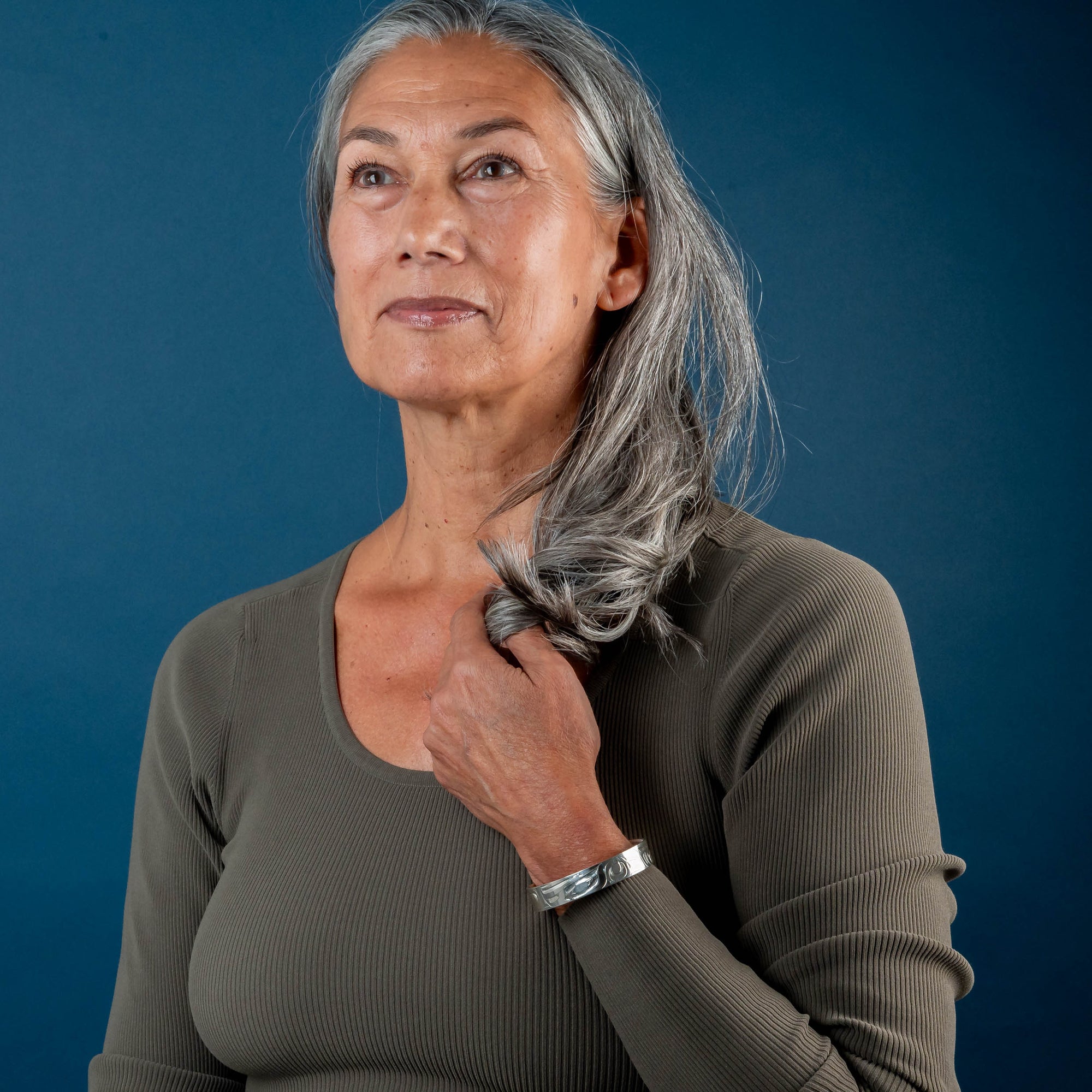 Woman with gray hair touching her shoulder against a blue background wearing a Hand carved sterling silver Eagle bracelet by Haida artist Alvin Adkins front view