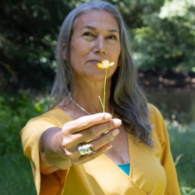 Woman holding a yellow flower outdoors with greenery in the background