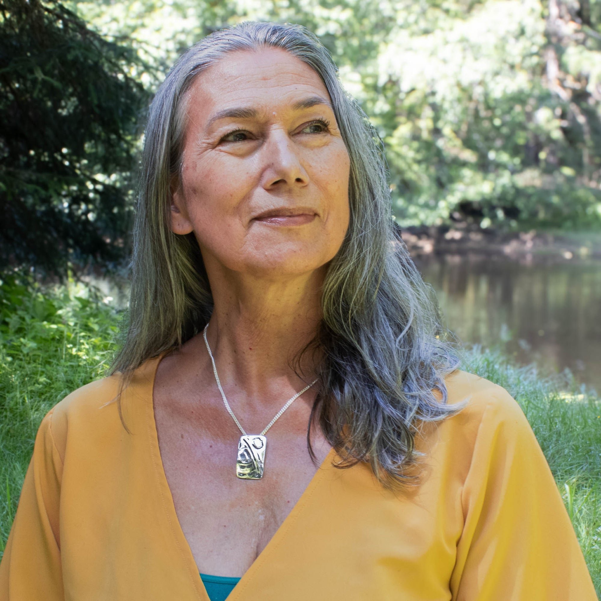 Woman with gray hair wearing a yellow top and  hummingbird necklace outdoors by a body of water.