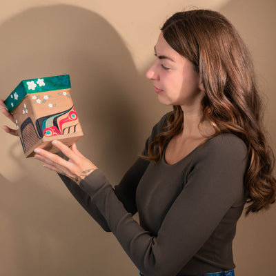 Woman holding a Haida Bentwood box with a colourful formline Beaver and floral formline design against a plain background