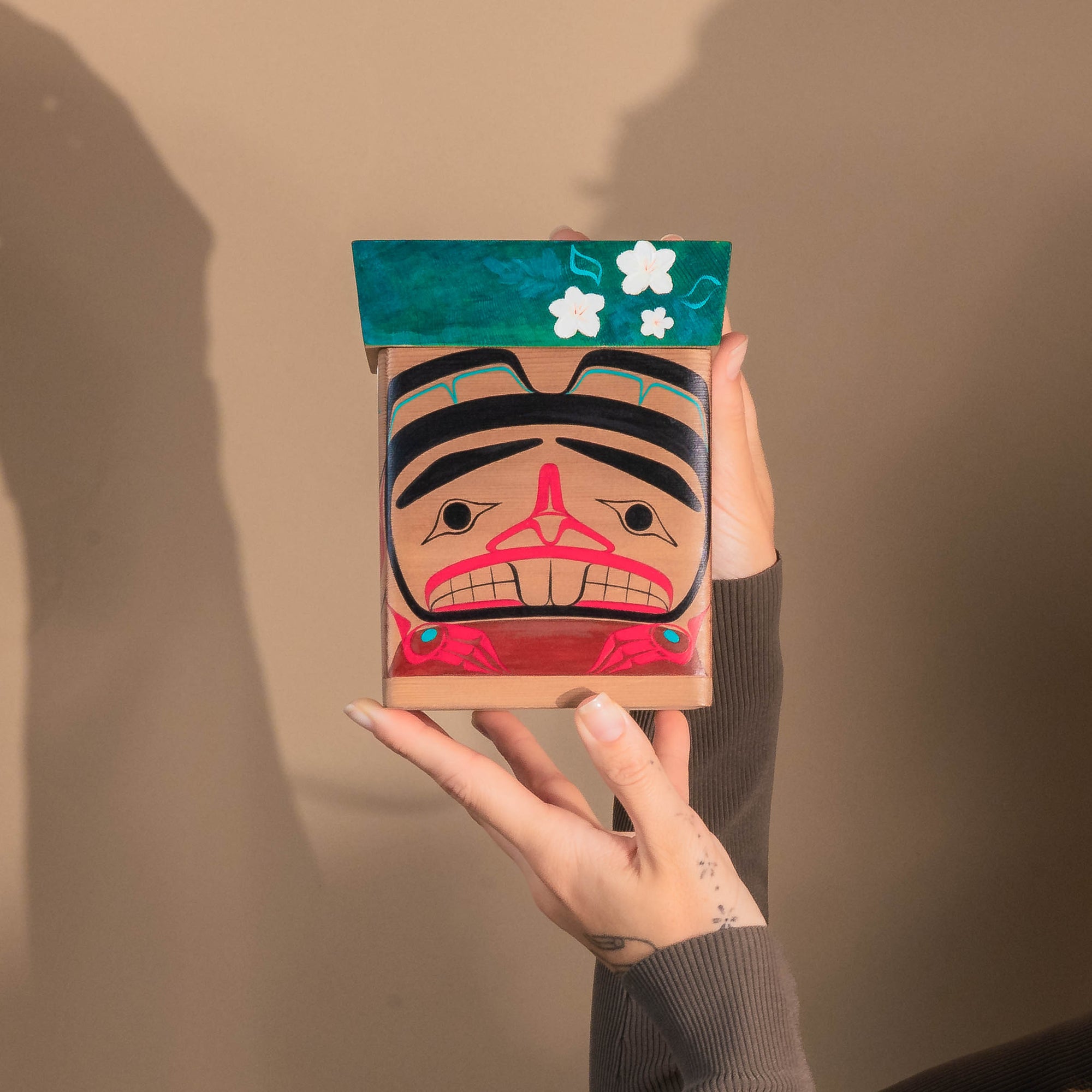 Hand holding a Haida Bentwood box with a colourful formline Beaver and floral formline design against a plain background