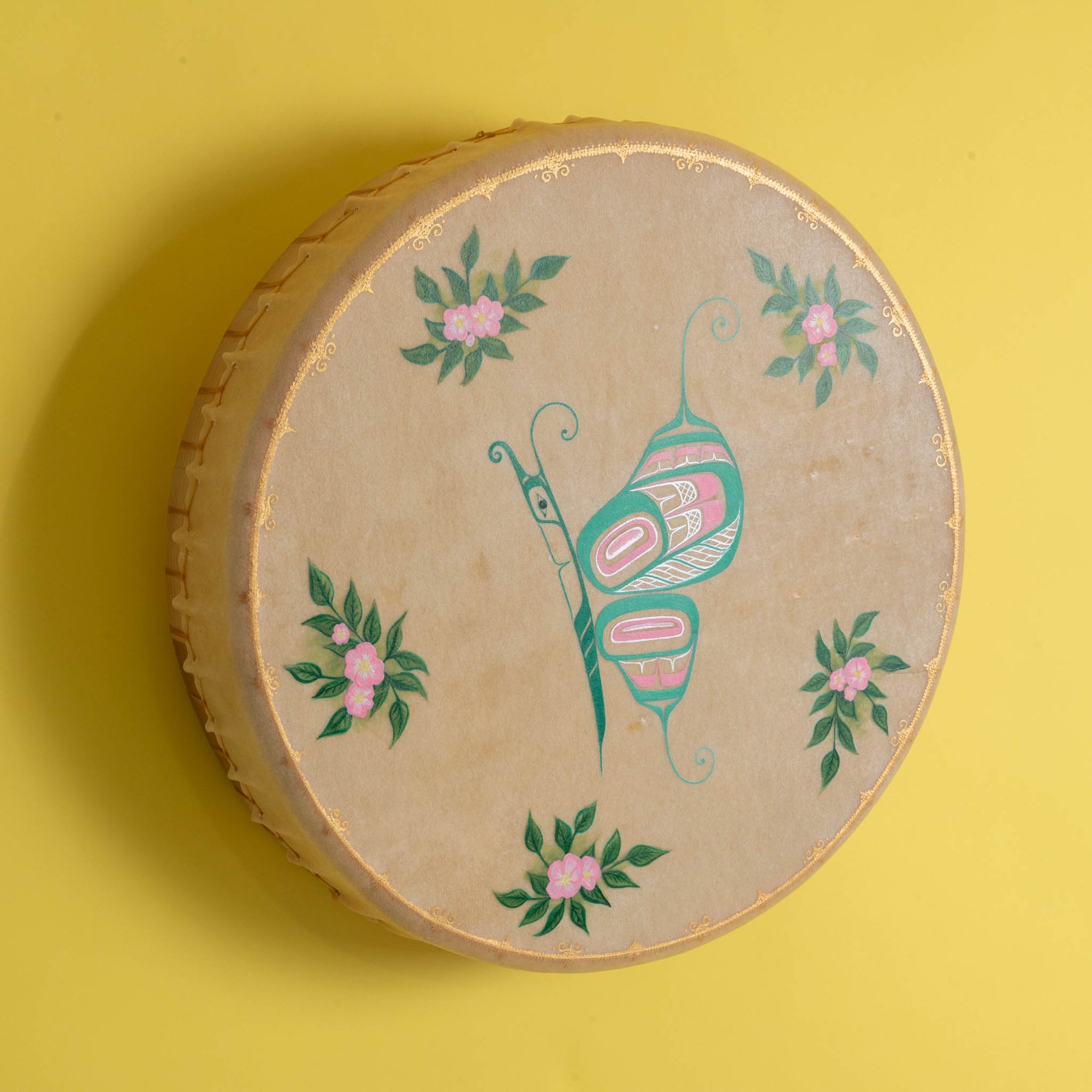 Tan drum with colourful shimmering butterfly in the center and flowers with a painted gold frame, on a yellow background.