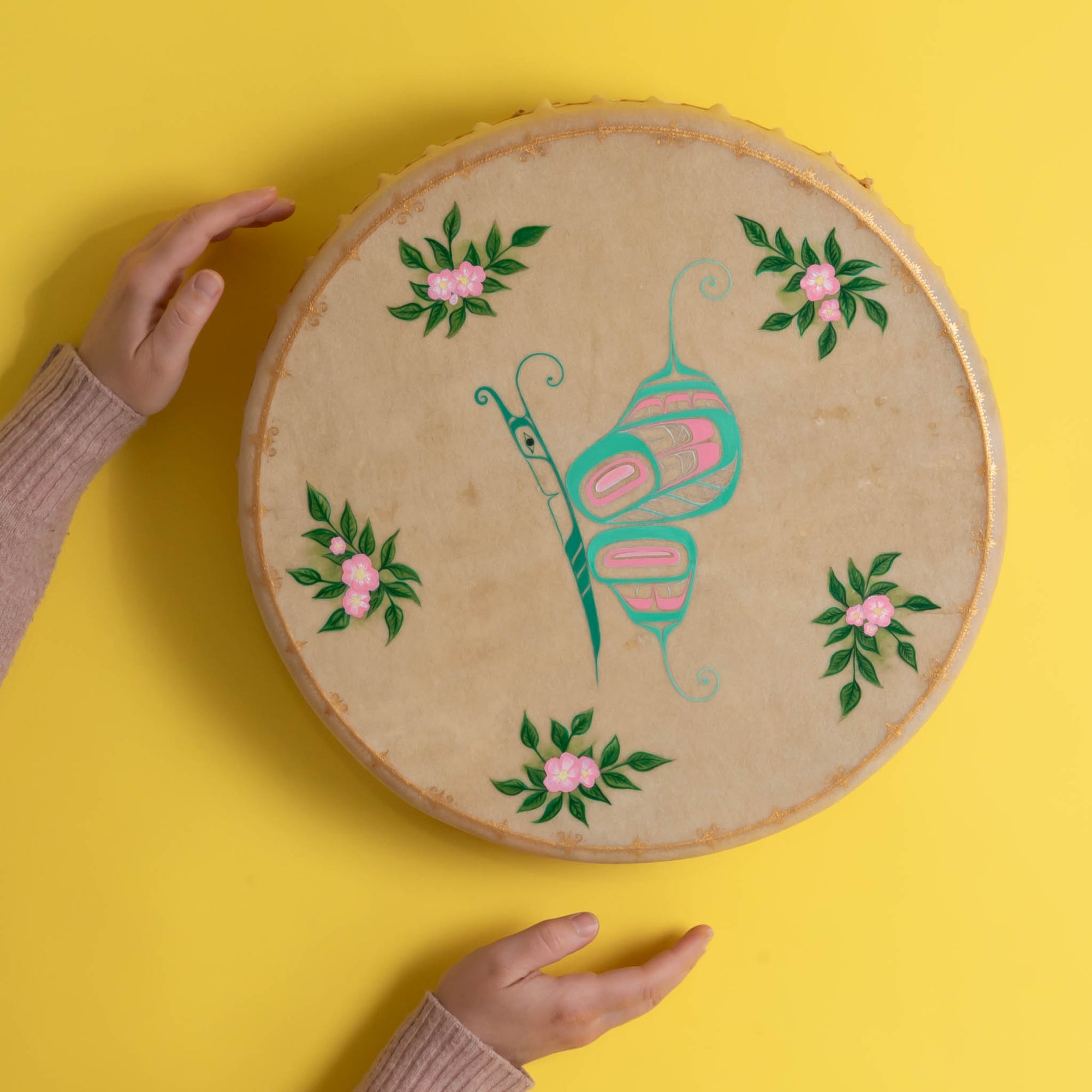 Tan drum with colourful butterfly in the center and flowers with a painted gold frame, on a yellow background with persons hands on left side.