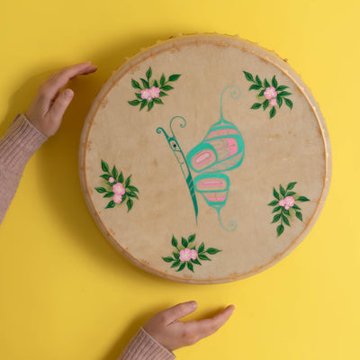 Tan drum with colourful butterfly in the center and flowers with a painted gold frame, on a yellow background with persons hands on left side.