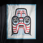 A blue, red, black and white Haida art print on a black background with a blue frame that has shadows reflecting on the glass of the art piece.