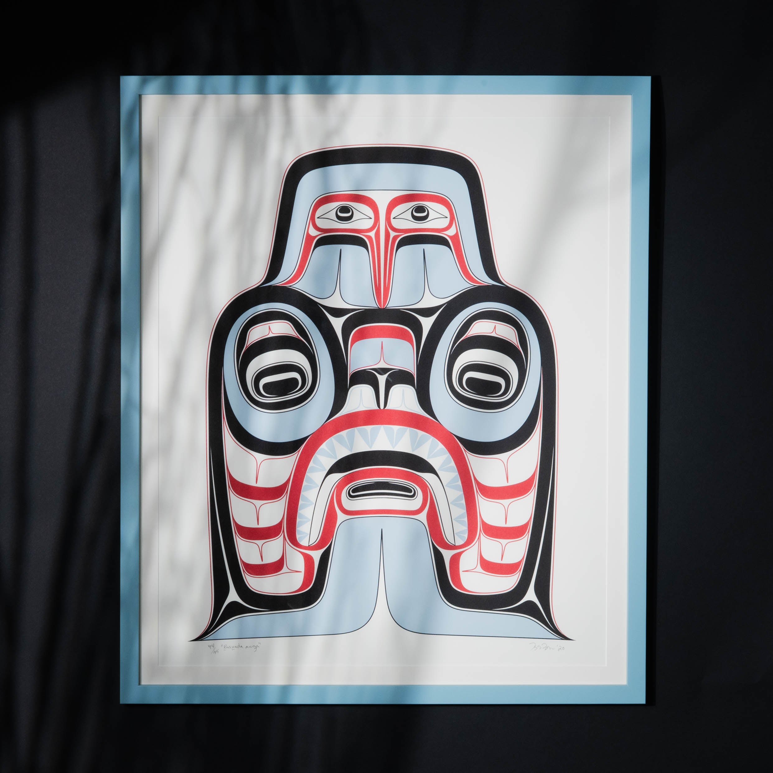 A blue, red, black and white Haida art print on a black background with a blue frame that has shadows reflecting on the glass of the art piece.
