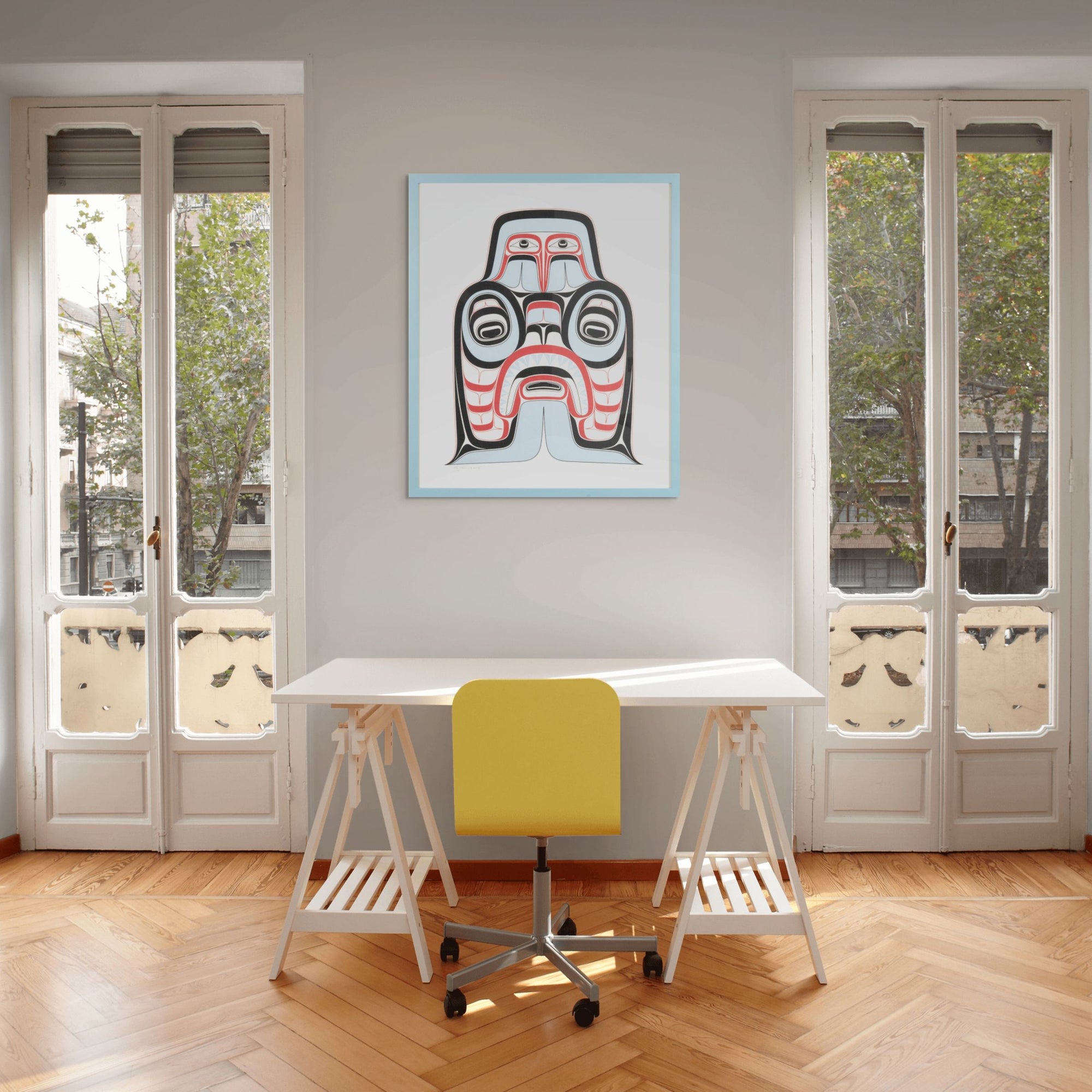 Modern office space with a white desk, yellow chair, and framed artwork of Haida Art print featuring supernatural being Dogfish mother. with bold red and black, and soft blue hues to show contrast, framed in a driftwood was frame on the wall.