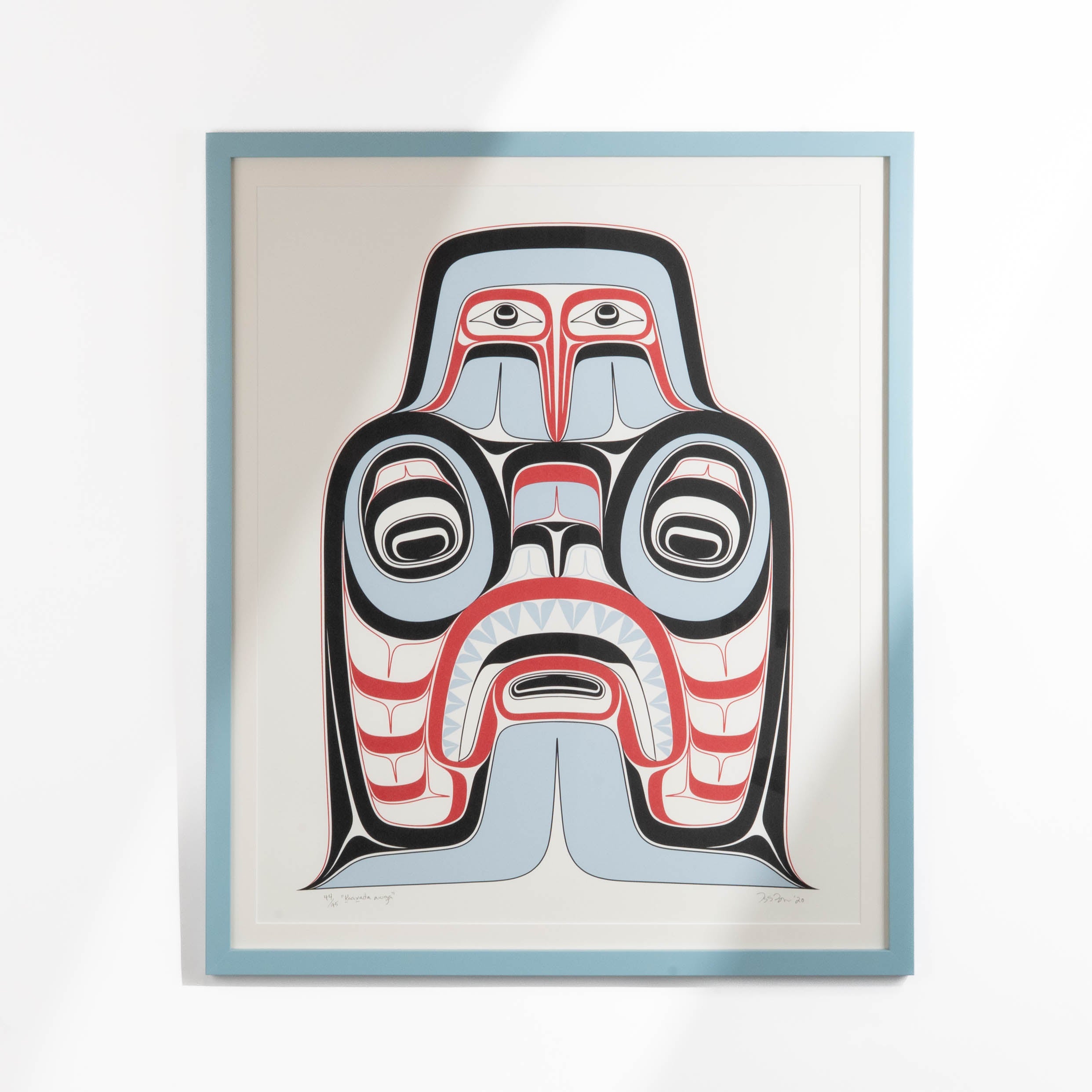 A white, red, black and light blue Haida northwest coast two-dimensional design framed in a blue frame.