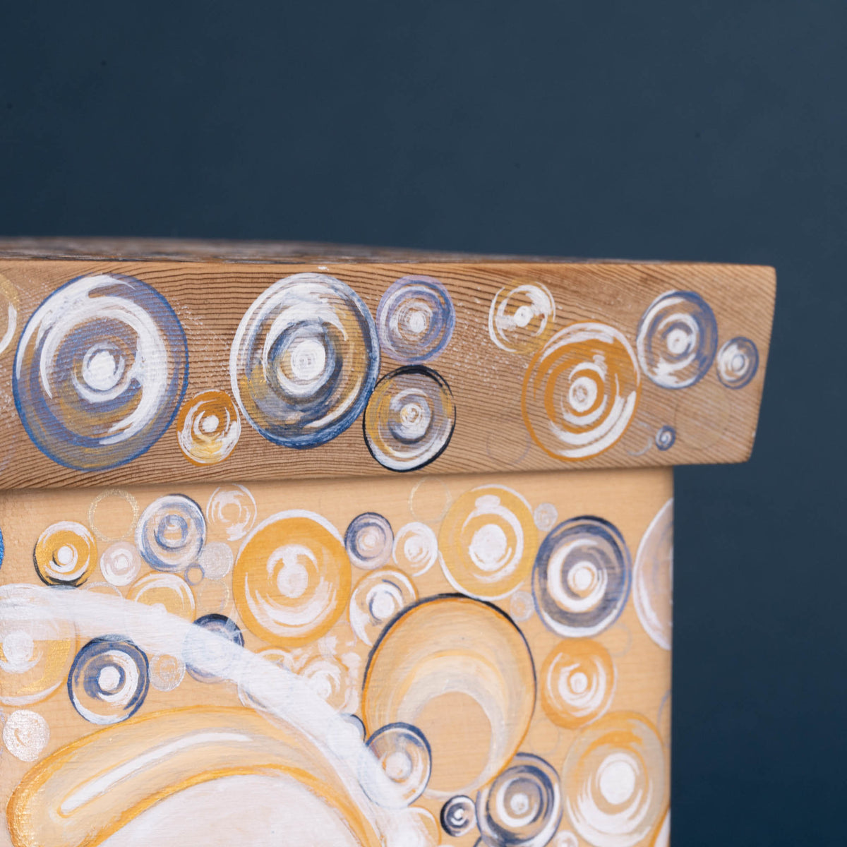 Close-up of a wooden box corner painted with circular bubble-like patterns in white, gray, blue, and warm yellow-orange tones against a dark blue background.