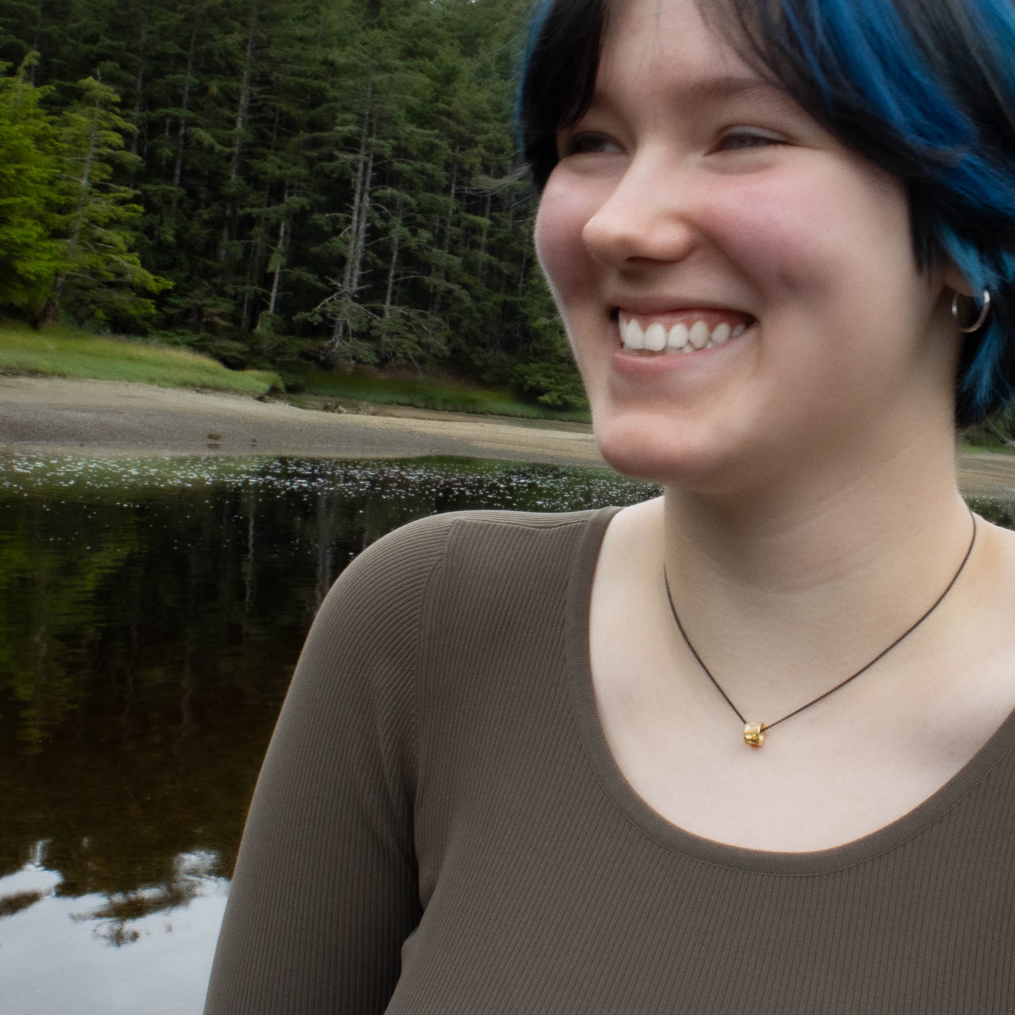 Person with blue hair smiling in front of a forest lake wearing a gold salmon bone vertebrae by Morgan Asoyuf