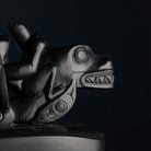 A close-up of a dark carved argillite depicting a killer whale with a person on its back, set against a very dark background.