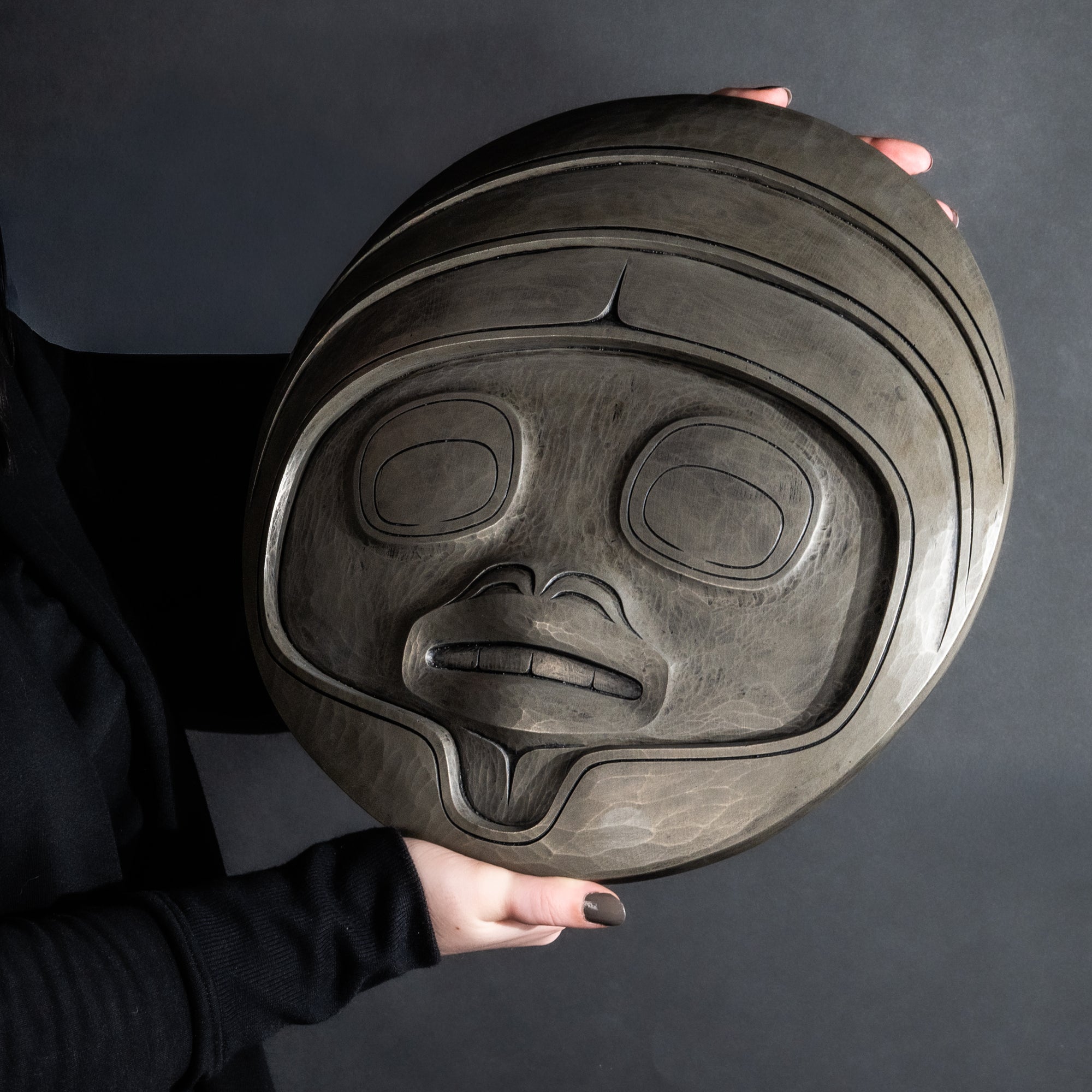 Person holding Fungus man mask casting with intricate carvings by Haida artist Cori Savard against a dark background