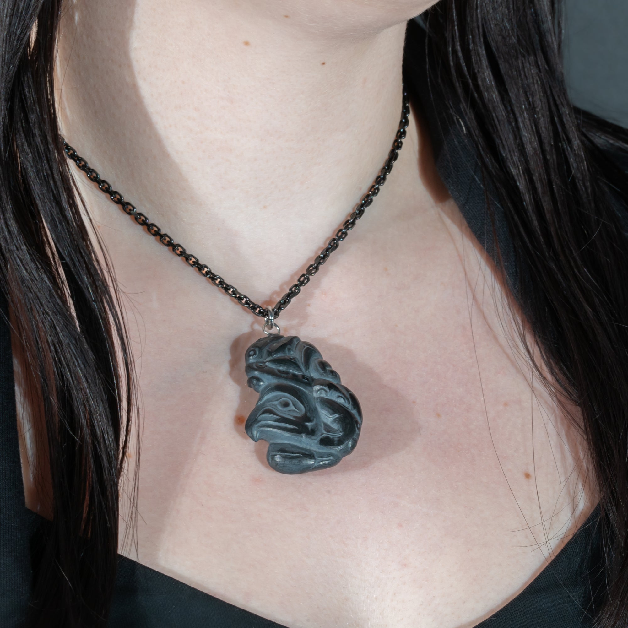 Close up of tentacles wrapping around both sides of Black Argillite Octopus pendant necklace with intricate Haida Carvings on a white background made by Local Haida Gwaii Artist Melanie Russ worn by a person with long hair.