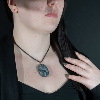 Person wearing a necklace Black Argillite Sea Bear pendant necklace with intricate Haida Carvings on a white background made by Local Haida Gwaii Artist Melanie Russ against a dark background