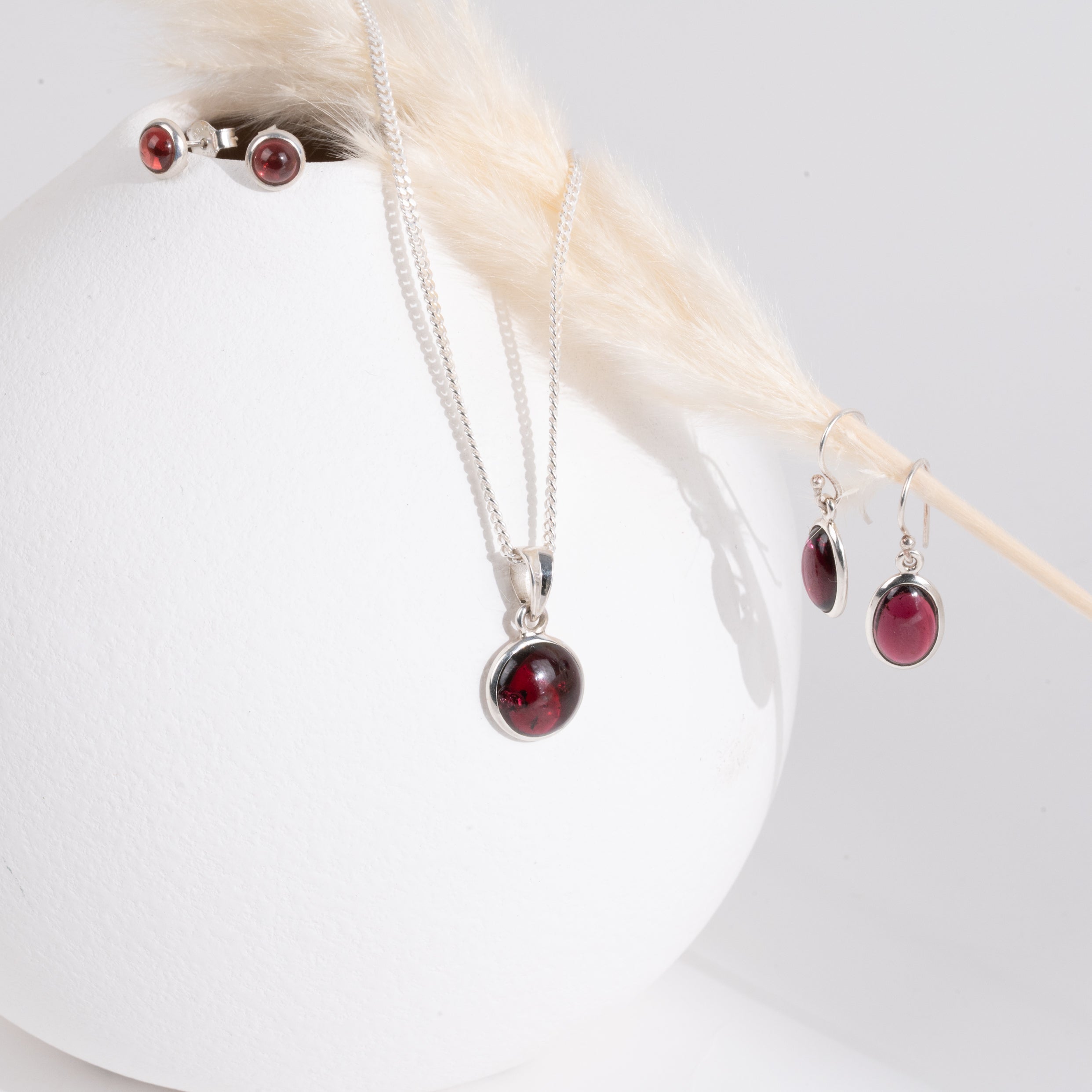 Jewelry set with red garnet gemstones by a white vase with a strand of yellow dried grass. 