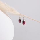 Pair of earrings with red garnet stones hanging on a strand of straw with a white vase on the left. 