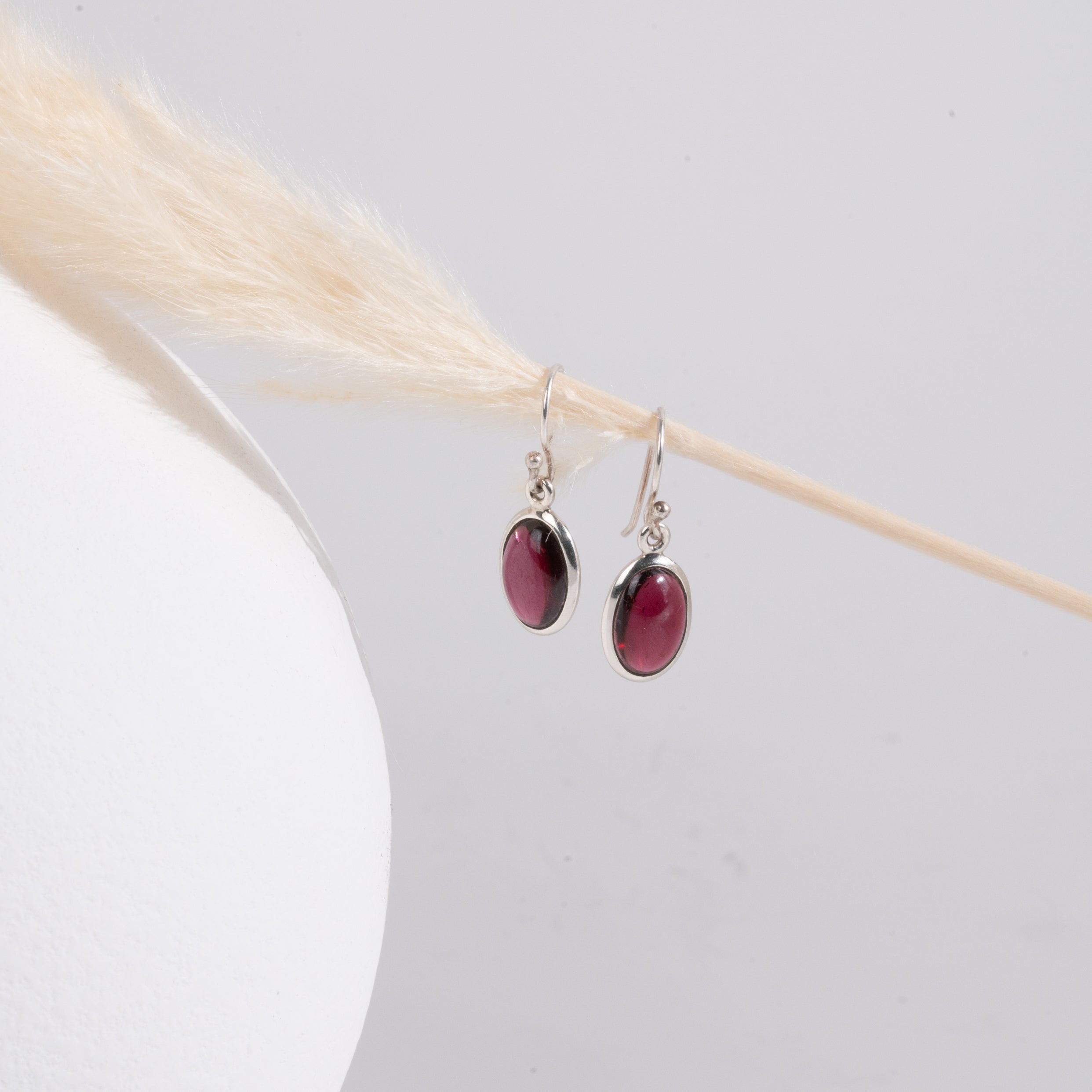 Pair of earrings with red garnet stones hanging on a strand of straw with a white vase on the left. 