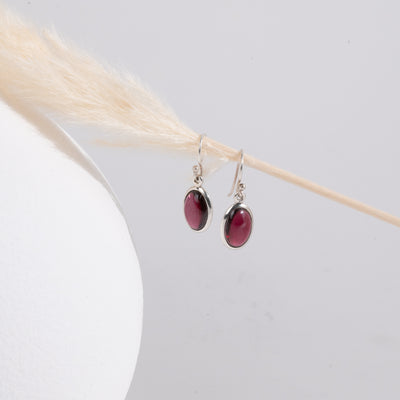 Pair of earrings with red garnet stones hanging on a strand of straw with a white vase on the left.