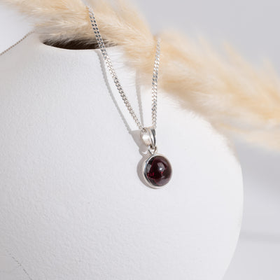 Silver necklace with a red garnet  gemstone pendant on a white surface with a piece of straw on the left.