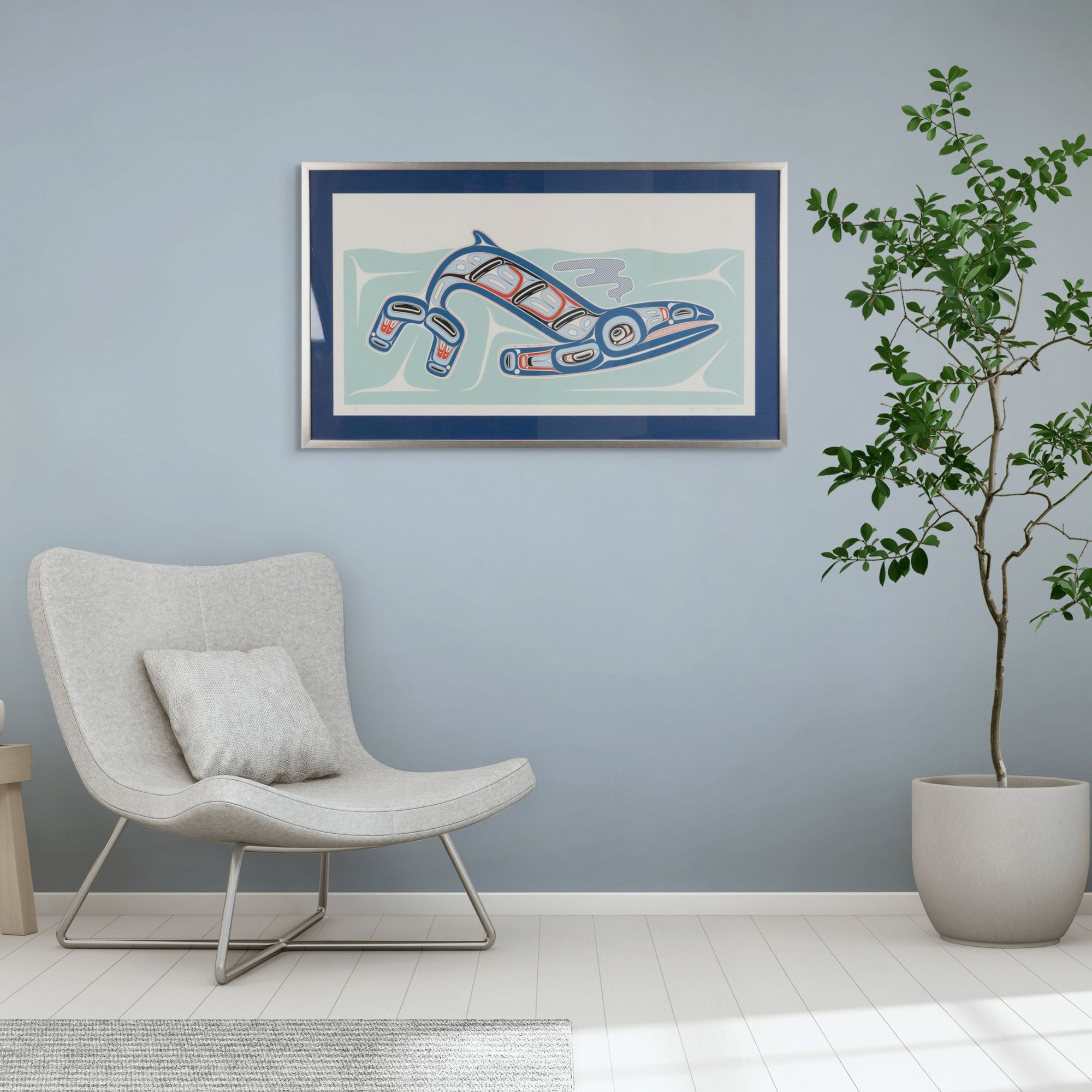 Modern living room with a chair, plant, and framed artwork of Kun (whale) Fine art print, showcasing a whale swimming through the ocean in Haida Formline. Set in an Interior designon a light blue wall.