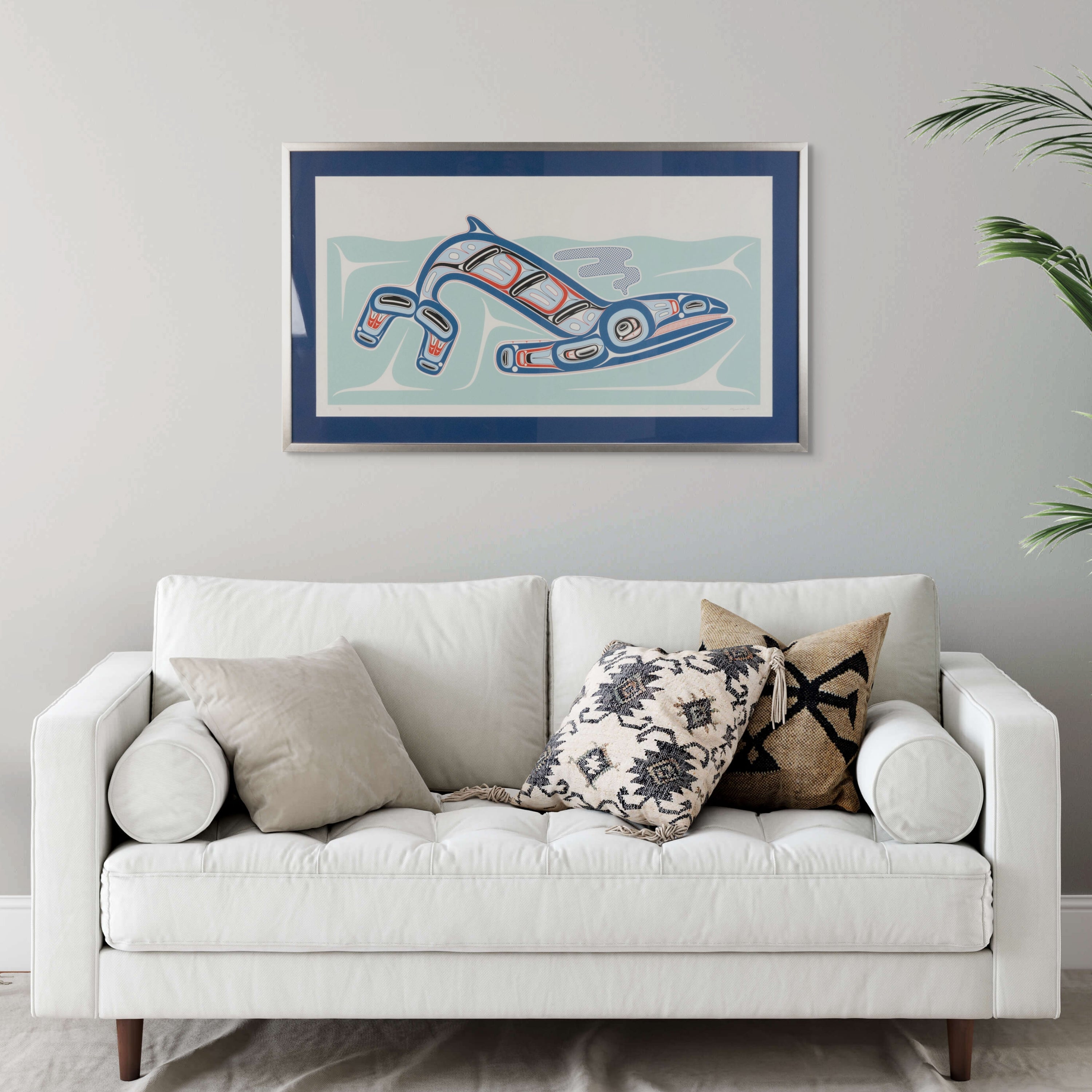 White sofa with decorative pillows in a room with a framed artwork of Kun (whale) Fine art print, showcasing a whale swimming through the ocean in Haida Formline on the wall.