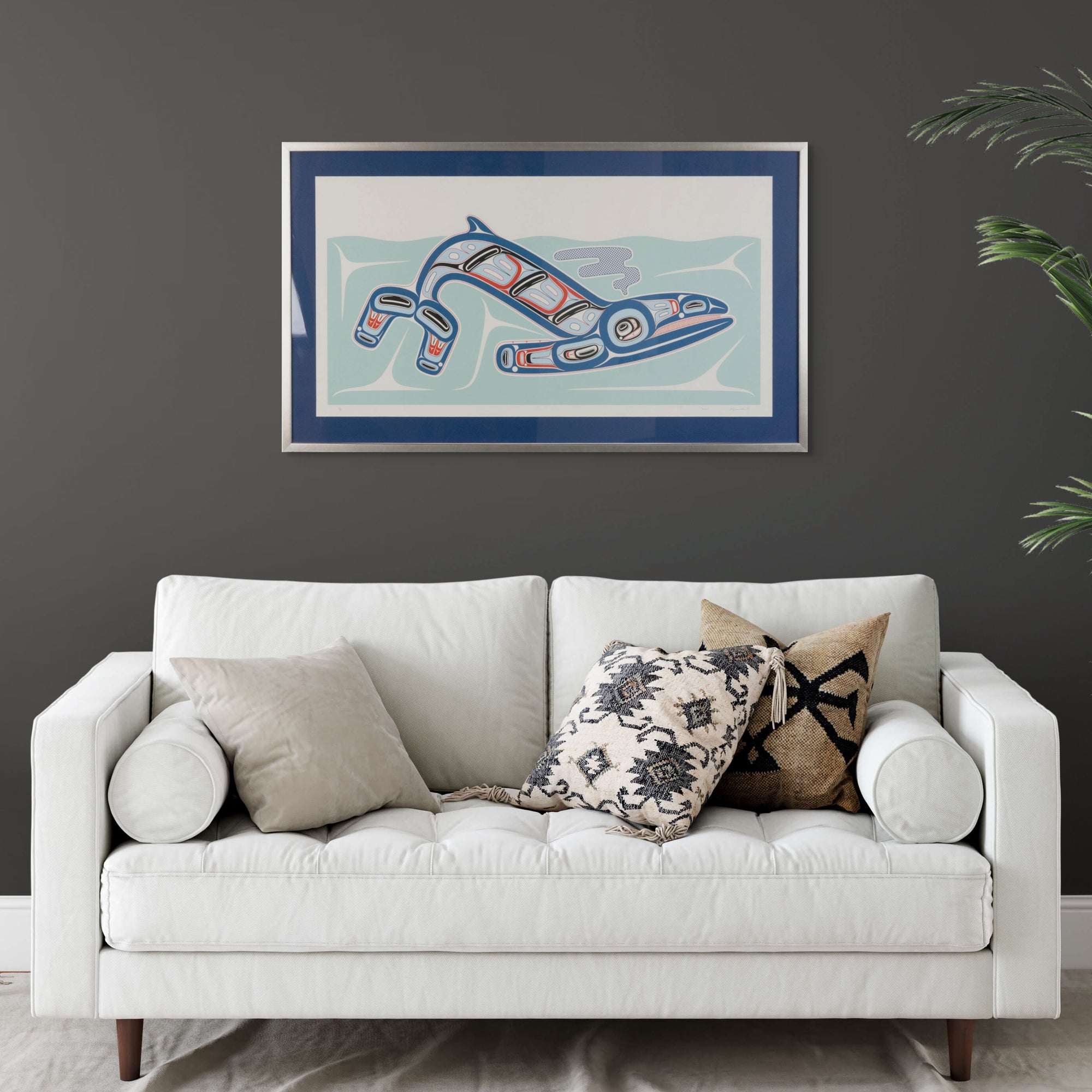 White sofa with decorative pillows against a dark gray wall with a framed abstract painting of Kun (whale) Fine art print, showcasing a whale swimming through the ocean in Haida Formline. Set in an Interior design