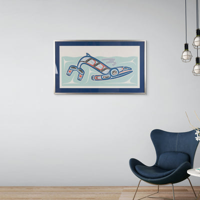 Framed artwork of Kun (whale) Fine art print, showcasing a whale swimming through the ocean in Haida Formline. Set in an Interior design on a wall above a modern blue chair.