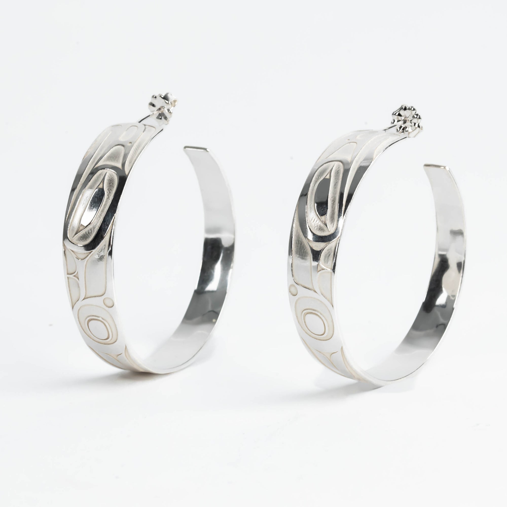 Pair of silver hoop earrings with intricate formline Ts'ymsen thunderbird designs on a white background