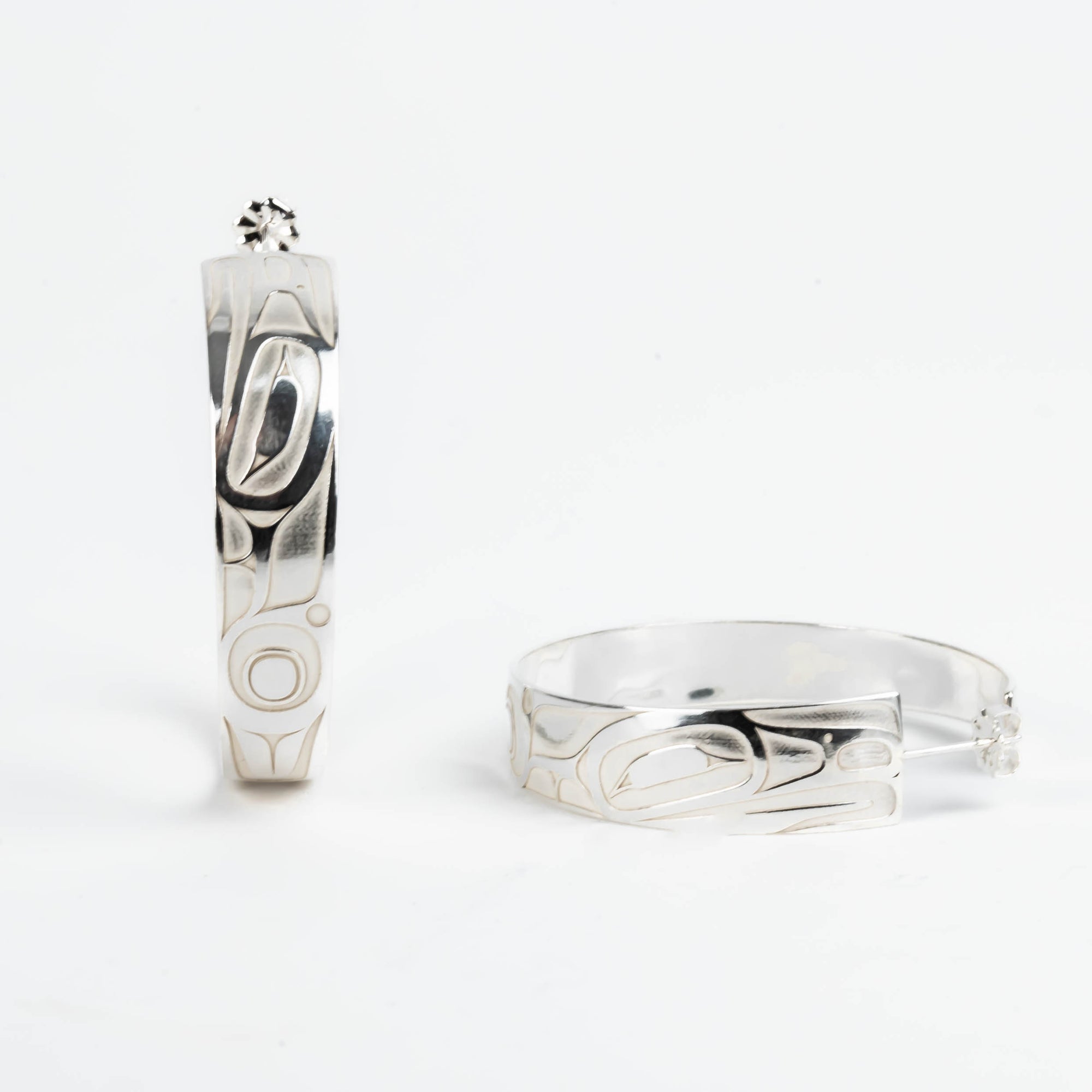 Pair of silver hoop earrings with intricate formline Ts'ymsen thunderbird designs on a white background