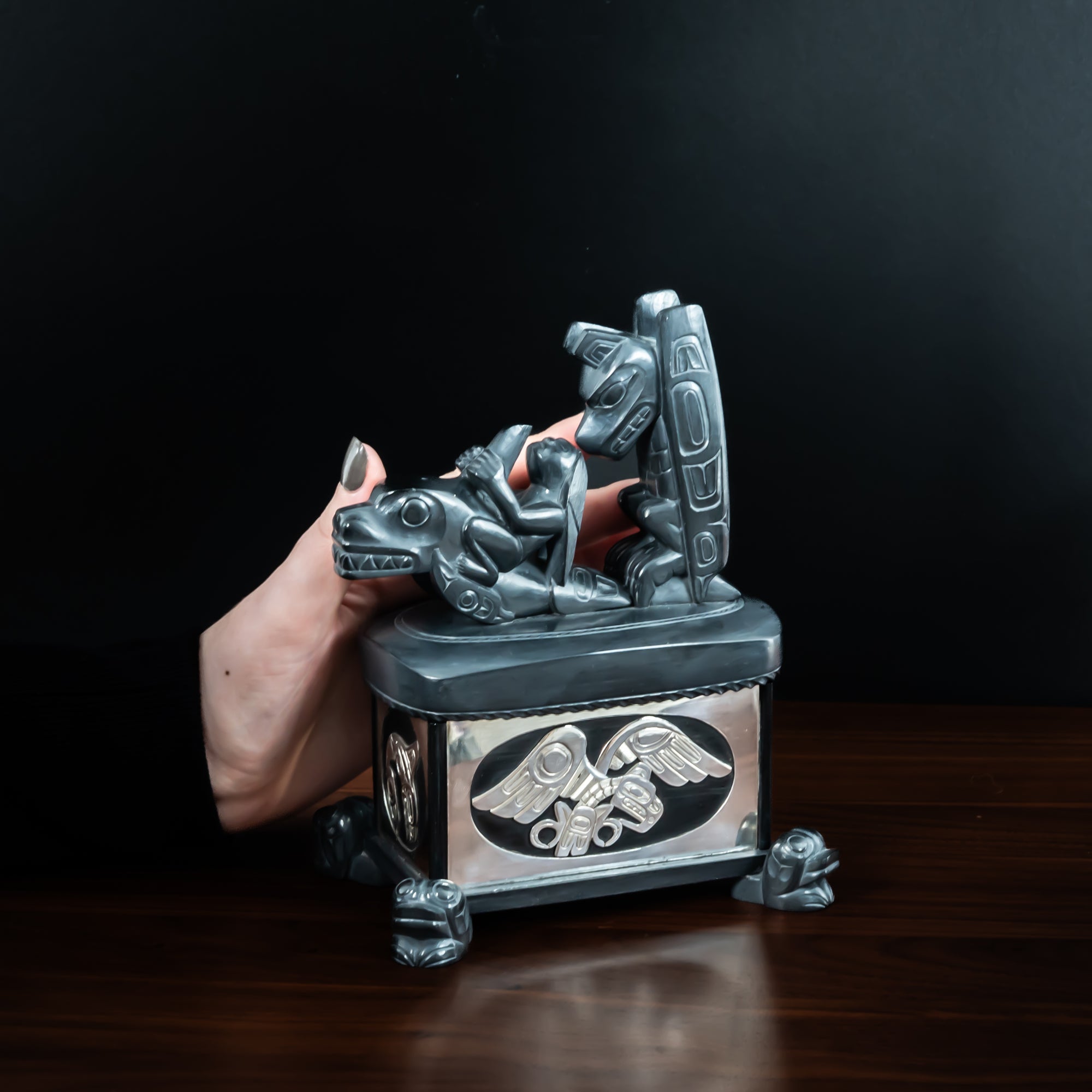 Hand touching Haida argillite killer whale and thunderbird argillite and sterling silver box done by Haida artist Ding Hutchingson on a dark background