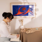Woman sitting at a table writing in a notebook with a colorful abstract painting of two dancing by Robert Davidson on the wall .