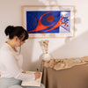 Woman sitting at a table writing in a notebook with a colorful abstract painting of two dancing by Robert Davidson on the wall .