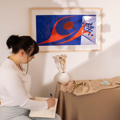 Woman sitting at a table writing in a notebook with a colorful abstract painting of two dancing by Robert Davidson on the wall .