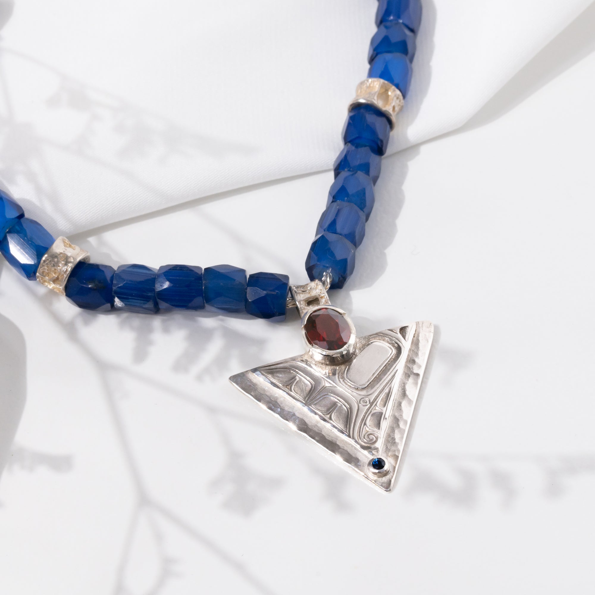 Necklace with Russian blue trade beads  and salmon bone vertebrae and a silver triangle hummingbird pendant with garnet and sapphire gems inlaid on a white background