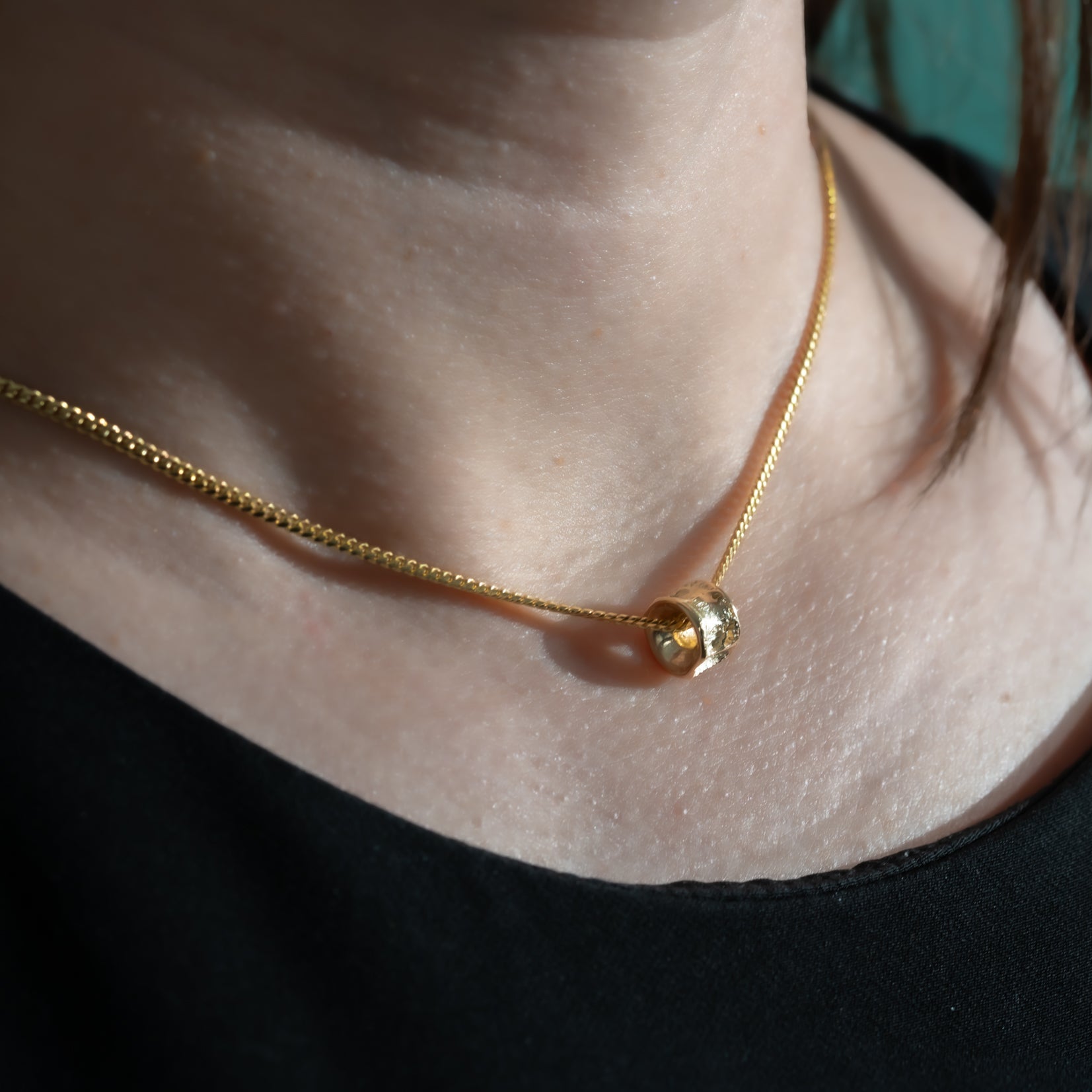 Gold necklace with a small round Salmon bone Vertebrae pendant on a person wearing a black top against a teal background